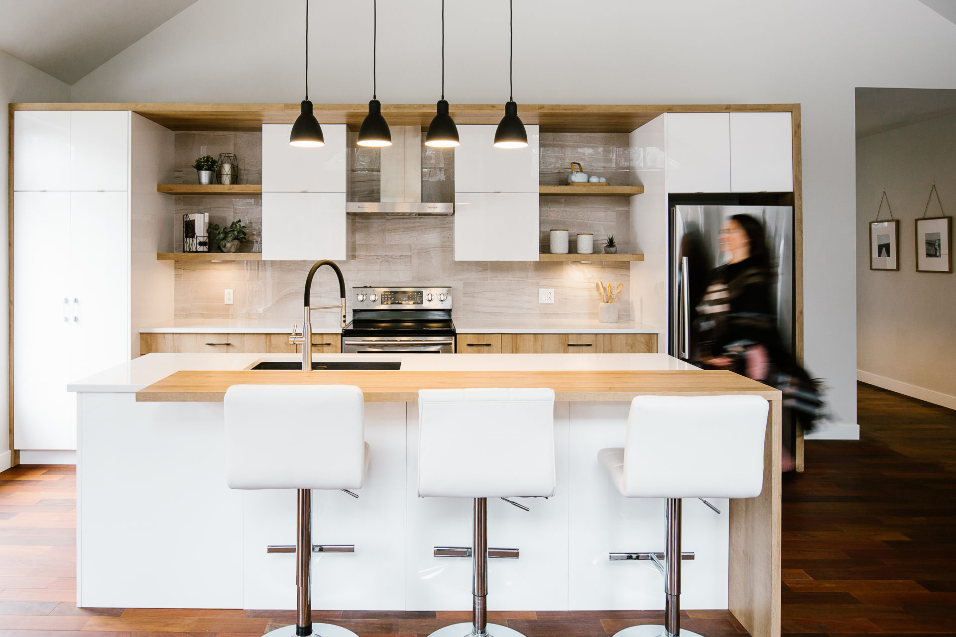 Cuisine blanche à îlot en bois, trois tabourets blancs et quatre suspensions noires.