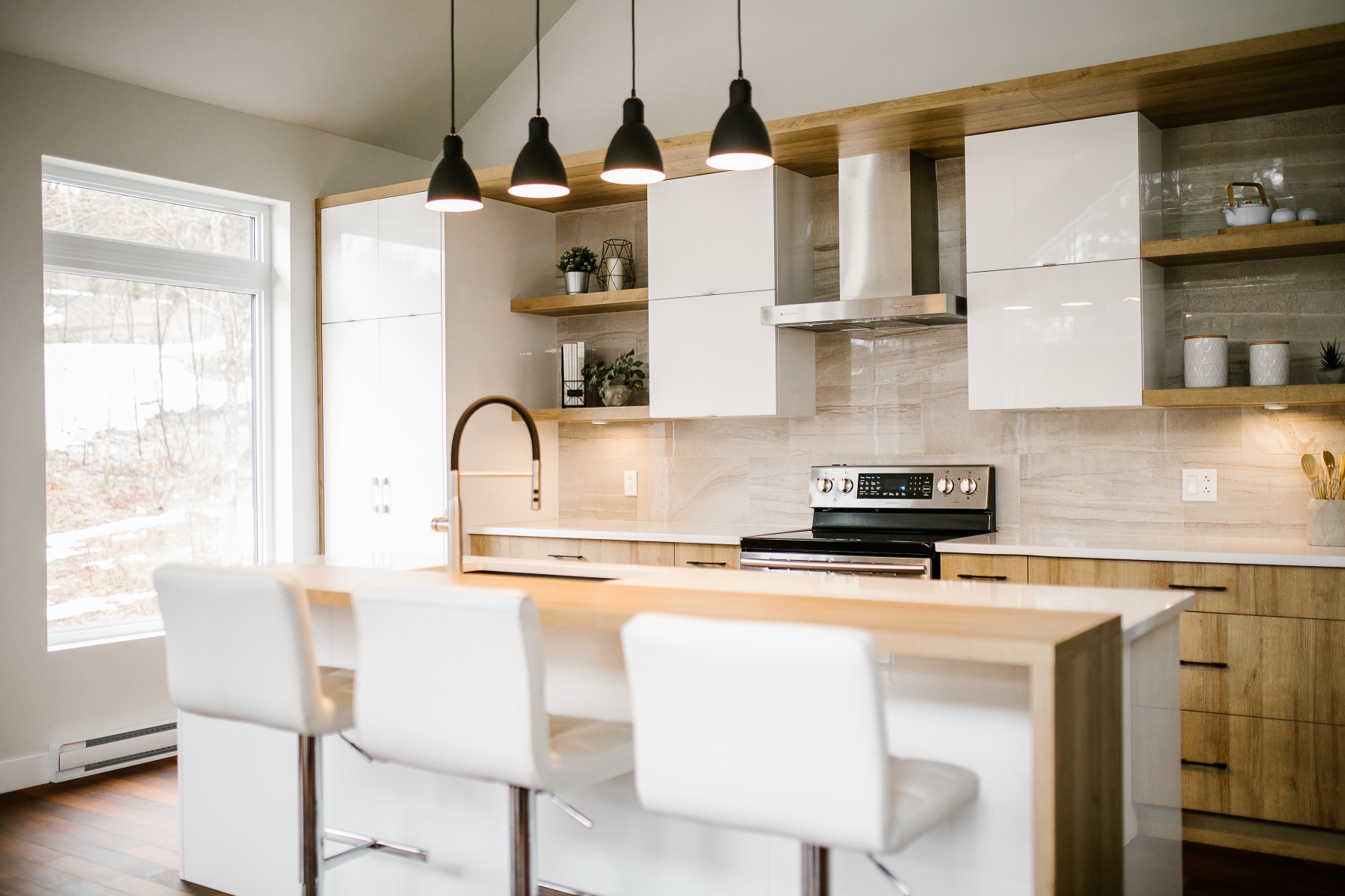 Cuisine blanche à îlot en bois clair, trois tabourets blancs et trois suspensions noires.