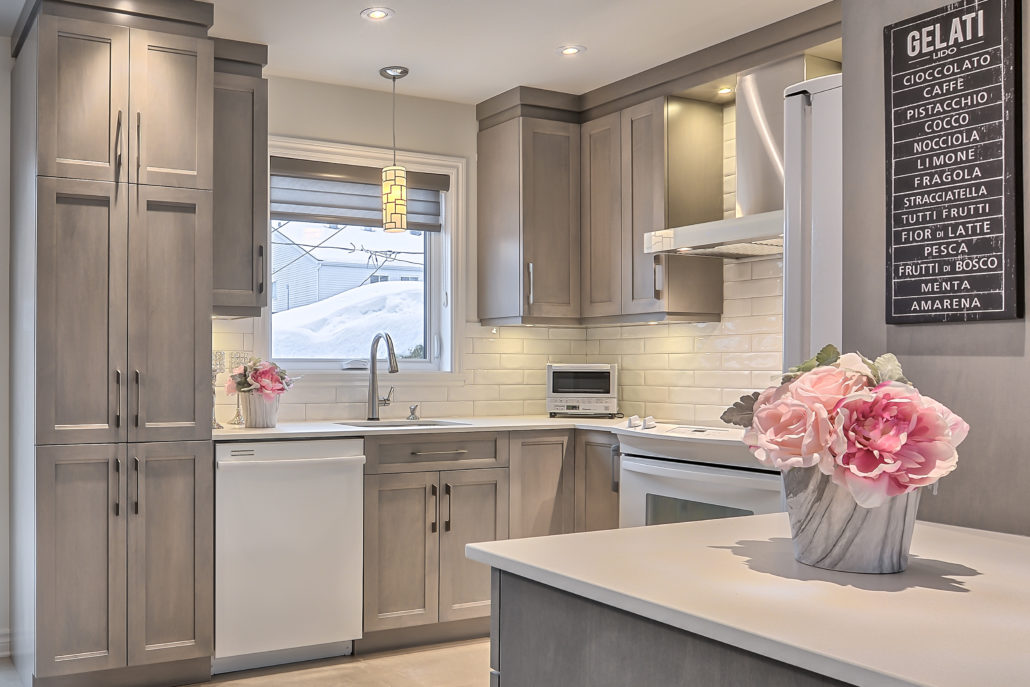 Cuisine aux armoires de bois teint gris, fleurs roses sur l’îlot et tableau « Gelati » au mur.
